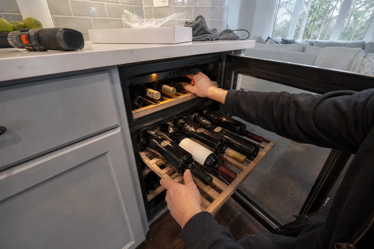 A hand slides a wooden rack of bottles out of an undercounter wine cooler with its glass door open, grey cabinets below and a cordless drill on the counter above